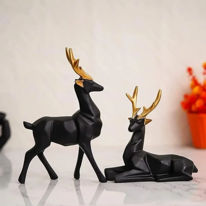 A set of two modern, low-poly geometric deer figurines with deep teal-green matte bodies and polished metallic gold antlers, positioned on a textured surface with a warm, blurred background of indoor plants.