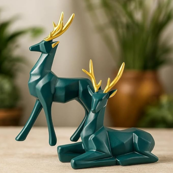 A set of two modern, low-poly geometric deer figurines with deep teal-green matte bodies and polished metallic gold antlers, positioned on a textured surface with a warm, blurred background of indoor plants.